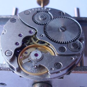 May include: Detailed view of a watch mechanism, featuring various gears, screws, and jewels. The silver metal parts are arranged in a circular design, revealing the internal workings of a watch. The image emphasizes the precision engineering of the timepiece.