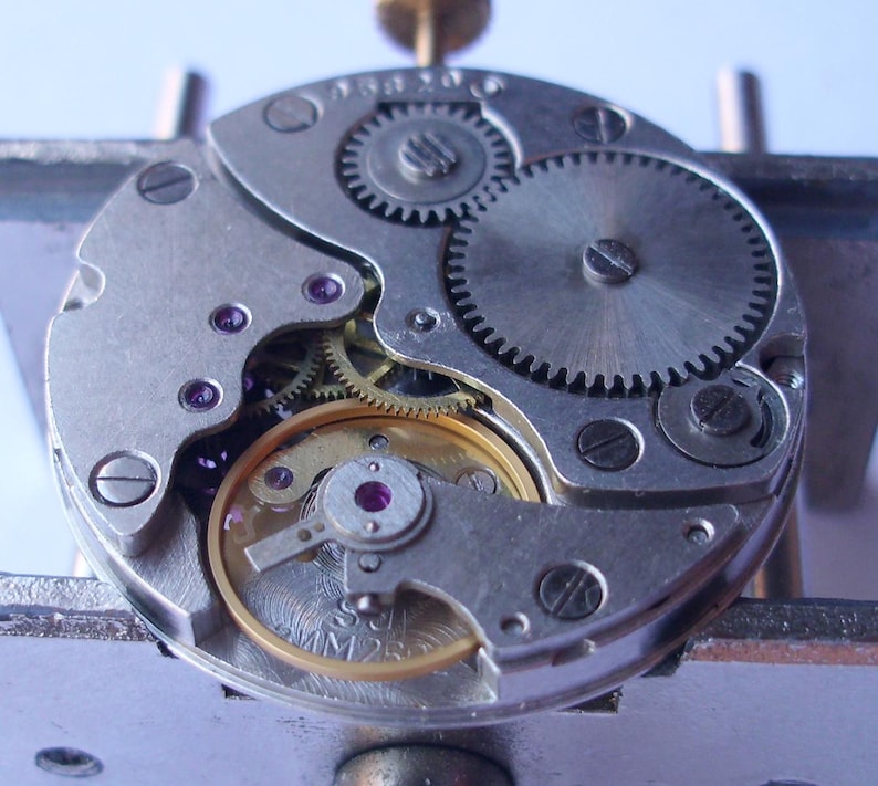 May include: Detailed view of a watch mechanism, featuring various gears, screws, and jewels. The silver metal parts are arranged in a circular design, revealing the internal workings of a watch. The image emphasizes the precision engineering of the timepiece.