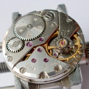 May include: A mechanical watch movement featuring a silver-toned main plate with circular graining, a gold-coloured balance wheel, and several purple synthetic rubies. The metal components include a gear with star engravings and text reading 18 jewels.