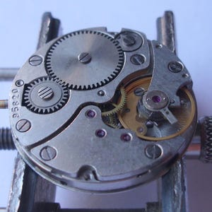 May include: Detailed view of a watch mechanism, showcasing intricate gears, screws, and internal parts. The metal components are silver and gold, with tiny red jewels. The number "95820" is visible on the watch movement.