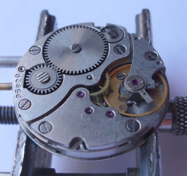 May include: Detailed view of a watch mechanism, showcasing intricate gears, screws, and internal parts. The metal components are silver and gold, with tiny red jewels. The number "95820" is visible on the watch movement.