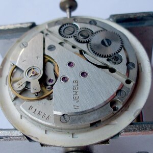 May include: Close-up of a watch movement with intricate gears, screws, and a balance wheel. The silver-colored mechanism features the text "17 JEWELS" and the number "01838". The watch is held in a metal clamp.