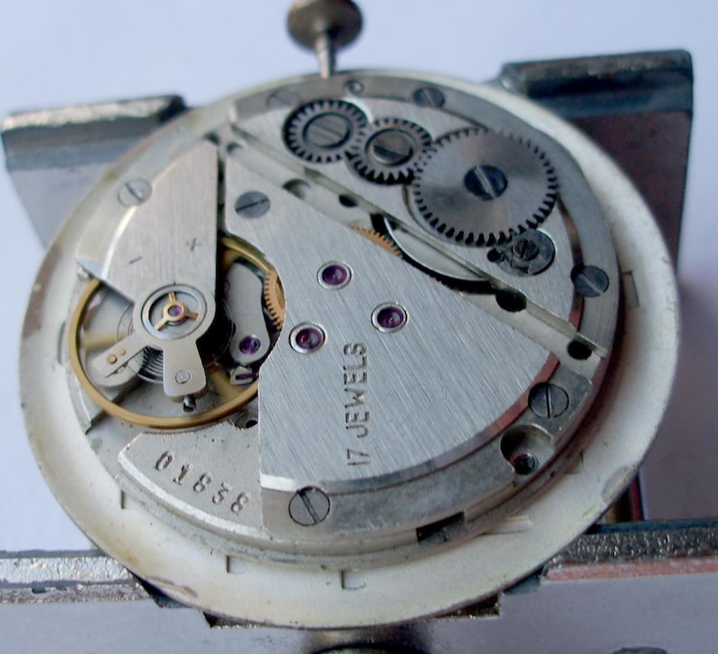 May include: Close-up of a watch movement with intricate gears, screws, and a balance wheel. The silver-colored mechanism features the text "17 JEWELS" and the number "01838". The watch is held in a metal clamp.