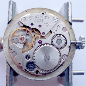 May include: Close-up of a watch movement with visible gears, springs, and jewels. The silver-toned mechanism is mounted in a holder. The text "16 KAMHEЙ 2603" is visible on the watch face.