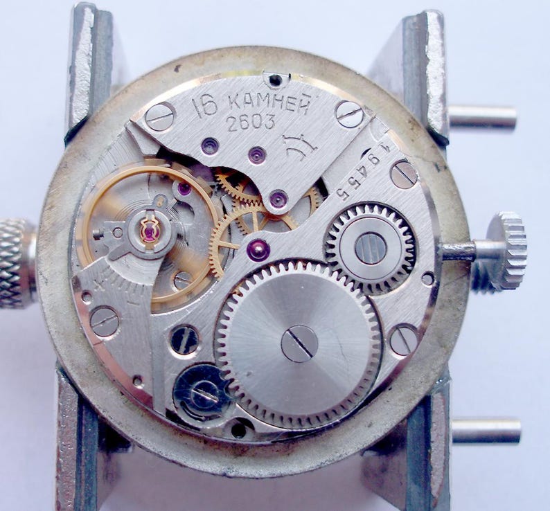 May include: Close-up of a watch movement with visible gears, springs, and jewels. The silver-toned mechanism is mounted in a holder. The text "16 KAMHEЙ 2603" is visible on the watch face.