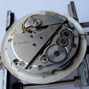 May include: Close-up of a watch movement with visible gears, screws, and the text "17 JEWELS". The watch mechanism is held in a metal clamp. The components are silver and gold-toned.