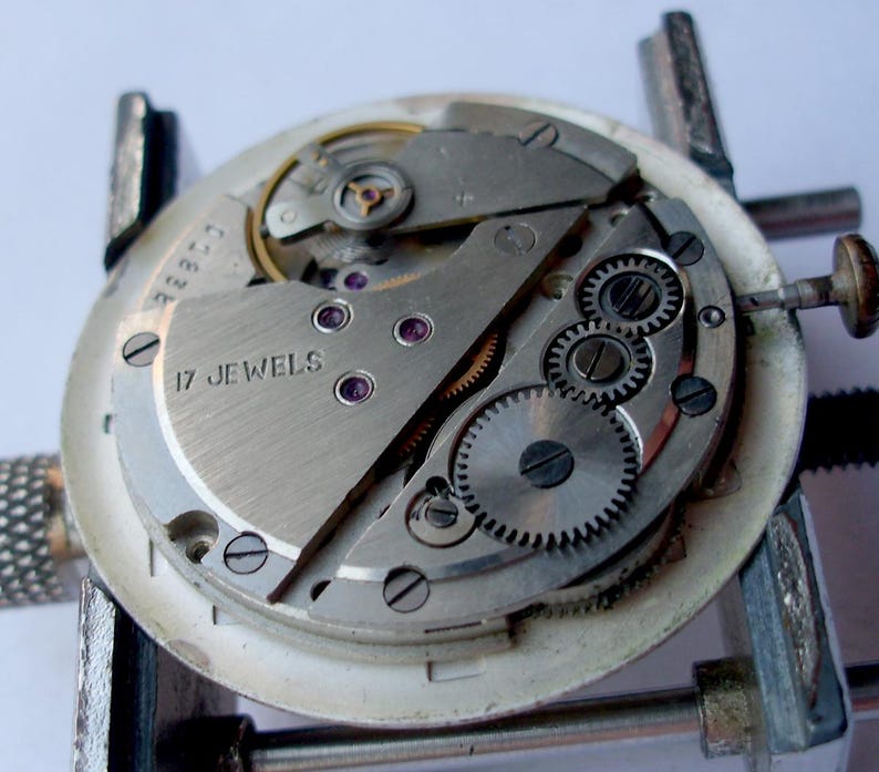 May include: Close-up of a watch movement with visible gears, screws, and the text "17 JEWELS". The watch mechanism is held in a metal clamp. The components are silver and gold-toned.