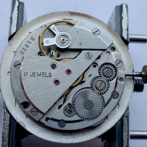 May include: Close-up of a watch movement with visible gears, screws, and the text "17 JEWELS". The silver-colored mechanism is mounted on a metal holder. The watch's internal components are exposed, showcasing intricate details.