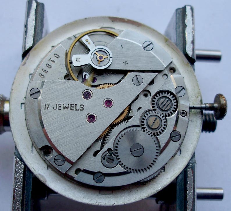 May include: Close-up of a watch movement with visible gears, screws, and the text "17 JEWELS". The silver-colored mechanism is mounted on a metal holder. The watch's internal components are exposed, showcasing intricate details.