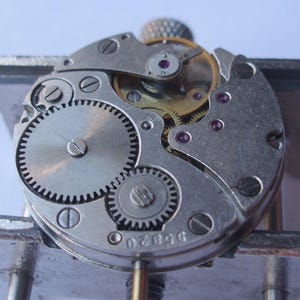 May include: Close-up of a watch mechanism showing intricate gears, screws, and jewels. The metal components are silver and gold, with small red jewels. The watch mechanism is held in a metal vice.
