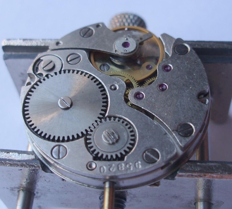 May include: Close-up of a watch mechanism showing intricate gears, screws, and jewels. The metal components are silver and gold, with small red jewels. The watch mechanism is held in a metal vice.