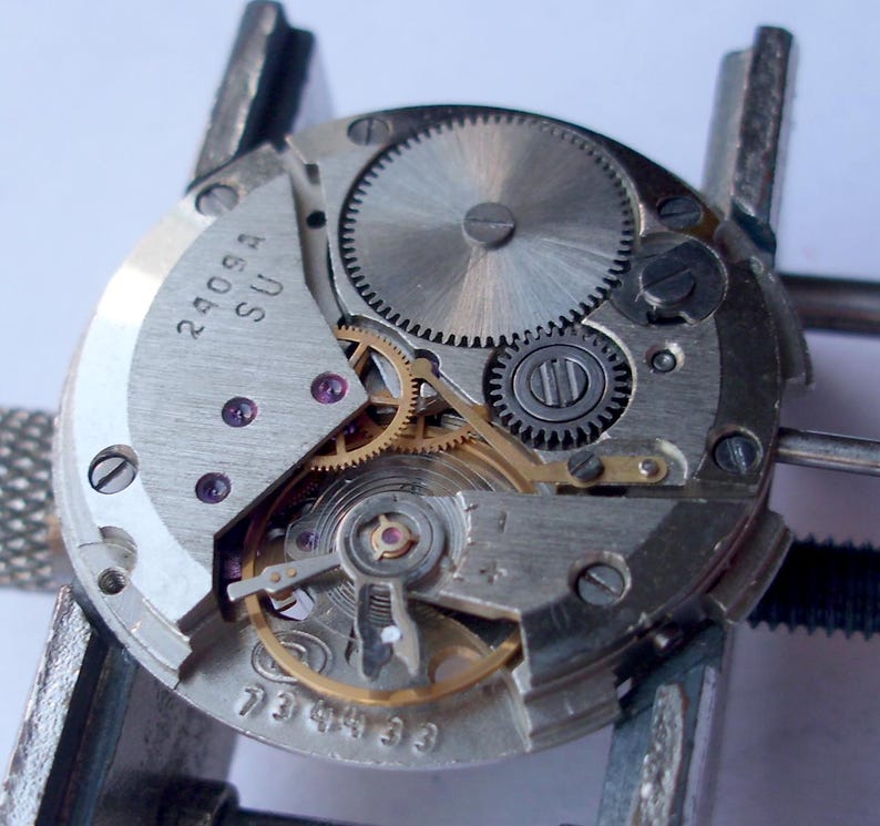 May include: Close-up of a watch movement with visible gears, screws, and other internal components. The metal parts are silver and gold, with small purple jewels. The text "2409A SU" and "734433" are visible on the movement.