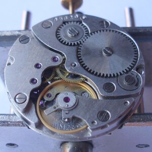 May include: Detailed view of a mechanical watch movement. The silver mechanism includes various gears, screws, and jewels. The intricate design reveals the internal workings of a timepiece, emphasizing its precision and craftsmanship.