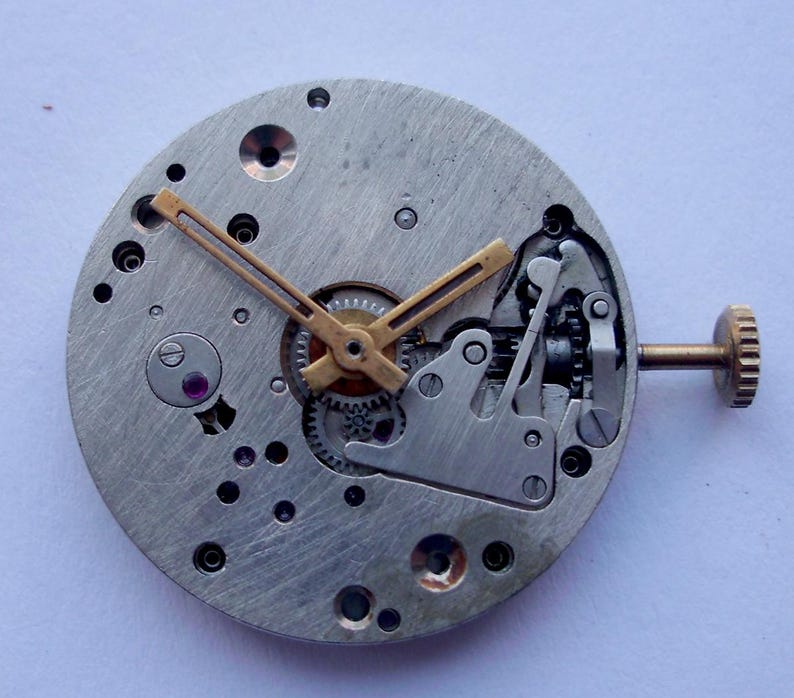 May include: Detailed view of a watch movement, revealing its internal components. The silver-coloured base houses gears, levers, and a gold-toned balance wheel. Gold-coloured hands indicate the time. A small, gold-coloured crown is present on the side.