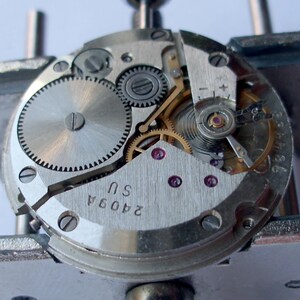 May include: Close-up of a watch movement with intricate gears, screws, and jewels. The silver-toned mechanism features a balance wheel, mainspring, and various other components. The text "2409A SU" is visible on the metal plate. The watch is held in place by a metal tool.