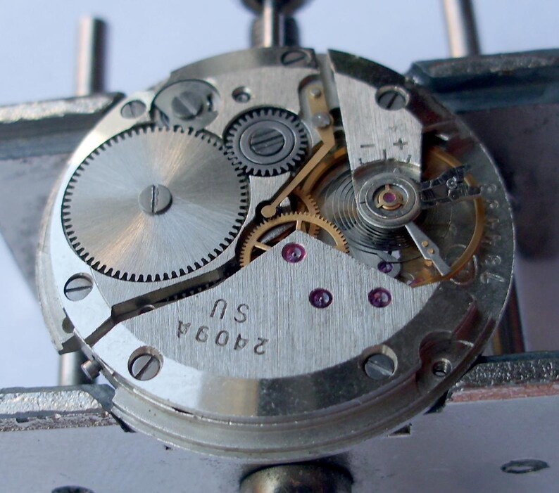May include: Close-up of a watch movement with intricate gears, screws, and jewels. The silver-toned mechanism features a balance wheel, mainspring, and various other components. The text "2409A SU" is visible on the metal plate. The watch is held in place by a metal tool.