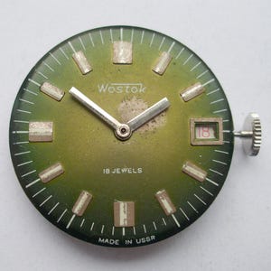 Pode incluir: Um mostrador de rel&oacute;gio vintage com mostrador verde, ponteiros prateados e marcadores de hora retangulares. O mostrador do rel&oacute;gio tem a marca "Wostok" e o texto "18 Jewels" e "Made in USSR". A data &eacute; vis&iacute;vel &agrave;s 3 horas.