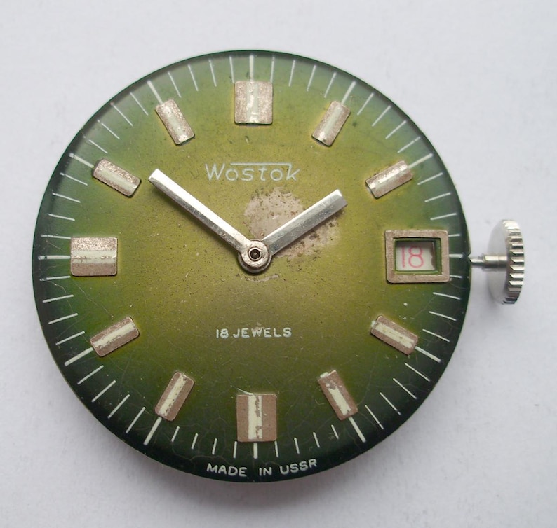 Pode incluir: Um mostrador de rel&oacute;gio vintage com mostrador verde, ponteiros prateados e marcadores de hora retangulares. O mostrador do rel&oacute;gio tem a marca "Wostok" e o texto "18 Jewels" e "Made in USSR". A data &eacute; vis&iacute;vel &agrave;s 3 horas.
