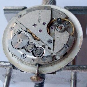 May include: Close-up of a watch movement with visible gears, screws, and other internal components. The watch mechanism is silver-toned with a white outer ring. The text "17 JEWELS" is visible.
