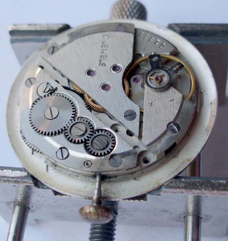 May include: Close-up of a watch movement with visible gears, screws, and other internal components. The watch mechanism is silver-toned with a white outer ring. The text "17 JEWELS" is visible.