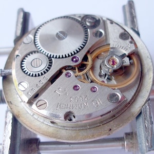 May include: Close-up of a watch movement with visible gears, screws, and jewels. The silver-colored mechanism is mounted on a metal holder. The text "16 KAMHEЙ 2603" is visible on the watch's face.