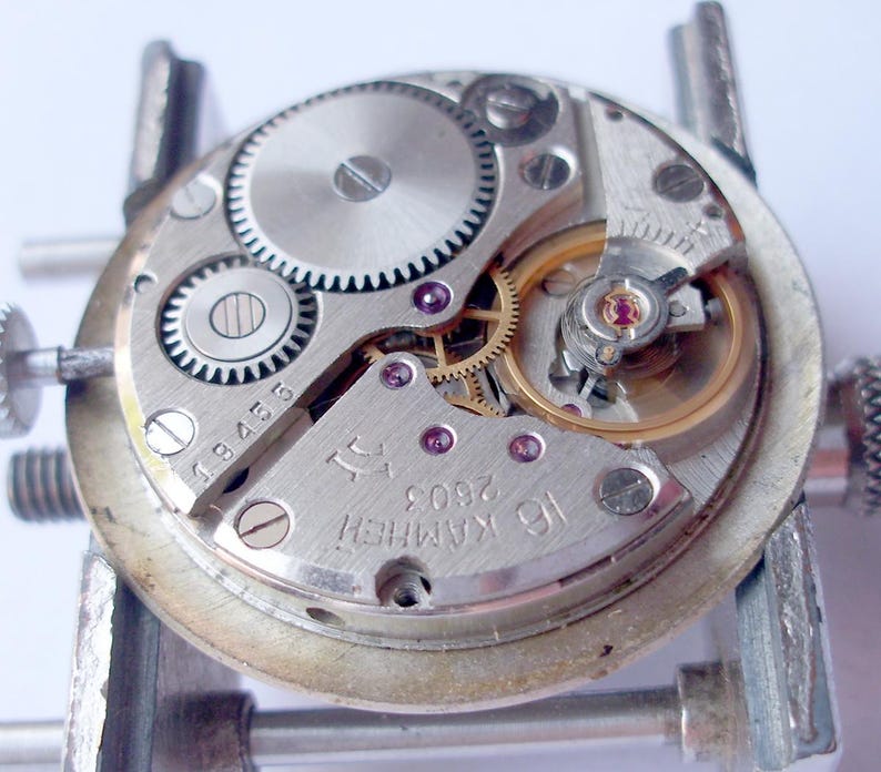 May include: Close-up of a watch movement with visible gears, screws, and jewels. The silver-colored mechanism is mounted on a metal holder. The text "16 KAMHEЙ 2603" is visible on the watch's face.