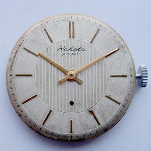 May include: A vintage Raketa watch face with a silver-colored dial and gold-tone hands and hour markers. The dial features a textured center and the Raketa brand name. The watch has a silver-colored case and a winding crown. The words "MADE IN USSR" are printed at the bottom.