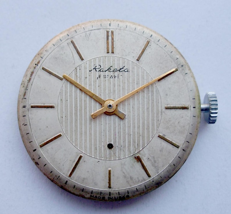 May include: A vintage Raketa watch face with a silver-colored dial and gold-tone hands and hour markers. The dial features a textured center and the Raketa brand name. The watch has a silver-colored case and a winding crown. The words "MADE IN USSR" are printed at the bottom.