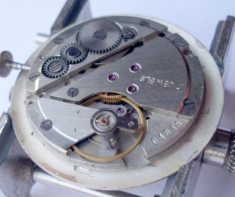 May include: Close-up of a watch movement with visible gears, screws, and jewels. The silver-toned components are arranged within a white circular frame, held by a metal clamp. The text "17 JEWELS" is visible.