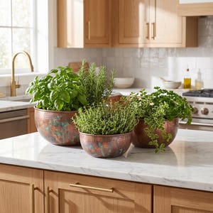 May include: Three round, hammered copper bowls with a weathered, teal-toned patina finish, each containing a different variety of fresh green herbs, including basil, rosemary, and thyme, arranged on a white marble kitchen worktop.