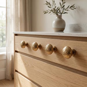 Puede incluir: Primer plano de una c&oacute;moda de madera clara con cuatro pomos ovalados de lat&oacute;n. Los pomos tienen un dise&ntilde;o floral detallado. Un jarr&oacute;n blanco con vegetaci&oacute;n se encuentra sobre la encimera encima de los cajones.