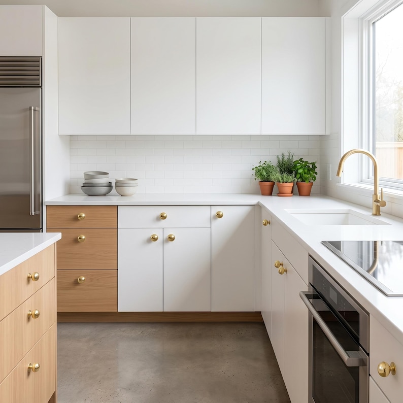 May include: Contemporary kitchen featuring white cabinets, a stainless steel fridge, and light wood drawers. White countertops and gold hardware are visible. Potted herbs are arranged on the countertop near a window.
