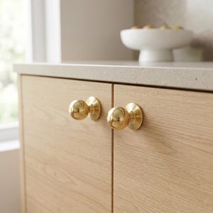 May include: Two sets of gold-coloured cabinet knobs are shown. The knobs are round with a circular base, mounted on light-coloured wooden cabinet doors. The knobs have a shiny, reflective surface. The cabinet doors have a natural wood grain.