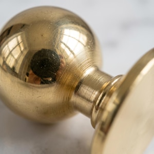 May include: Close-up of a brass seal stamp. The stamp features a round, polished base, a cylindrical neck, and a spherical top. The brass surface reflects light, creating a warm, golden hue. The stamp is designed with detailed, concentric circular patterns.