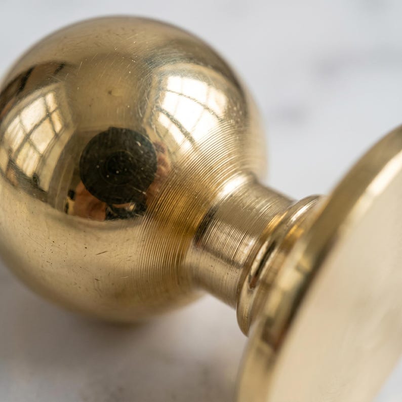 May include: Close-up of a brass seal stamp. The stamp features a round, polished base, a cylindrical neck, and a spherical top. The brass surface reflects light, creating a warm, golden hue. The stamp is designed with detailed, concentric circular patterns.