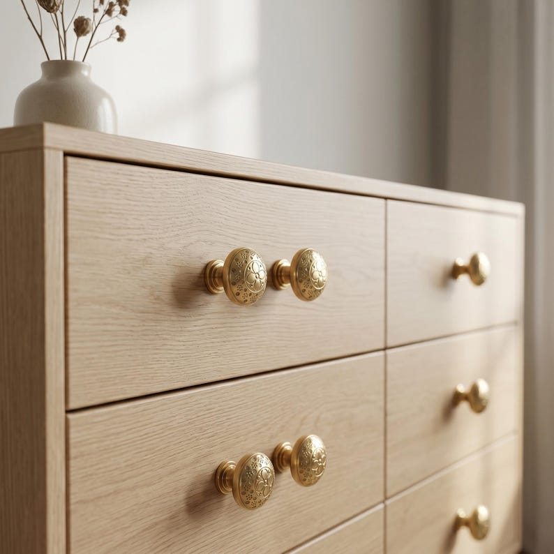 Puede incluir: Una c&oacute;moda de madera clara con seis cajones, cada uno con tiradores dorados ornamentados. Los tiradores tienen un dise&ntilde;o en relieve detallado. Un peque&ntilde;o jarr&oacute;n con flores secas se encuentra encima de la c&oacute;moda.