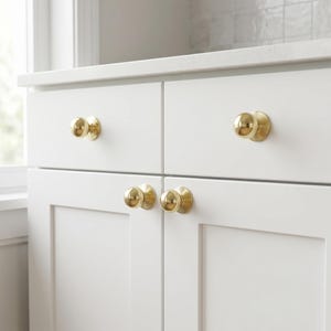 May include: A set of white cabinet doors with four gold-coloured, round cabinet knobs. The knobs are evenly spaced on the doors, which are part of a white cabinet set. The cabinet is set against a white countertop and a light-coloured tiled backsplash.