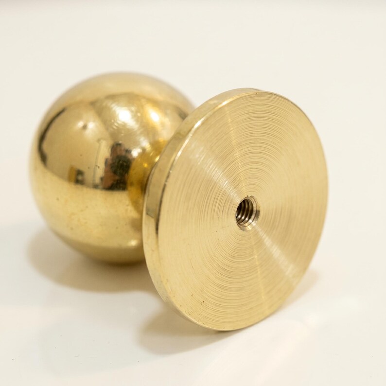 May include: A gold-coloured metal furniture knob with a spherical top and a flat, circular base. The base has a threaded hole in the centre. The knob is designed for attaching to furniture.