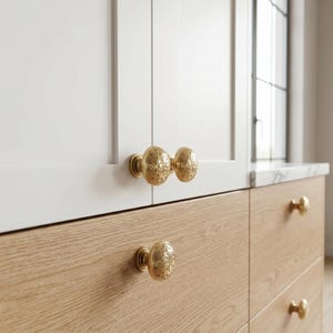 Puede incluir: Primer plano de gabinetes blancos y de madera con pomos de gabinete ovalados dorados y ornamentados. Los pomos tienen un dise&ntilde;o floral detallado. Los gabinetes est&aacute;n en un entorno de cocina o ba&ntilde;o.
