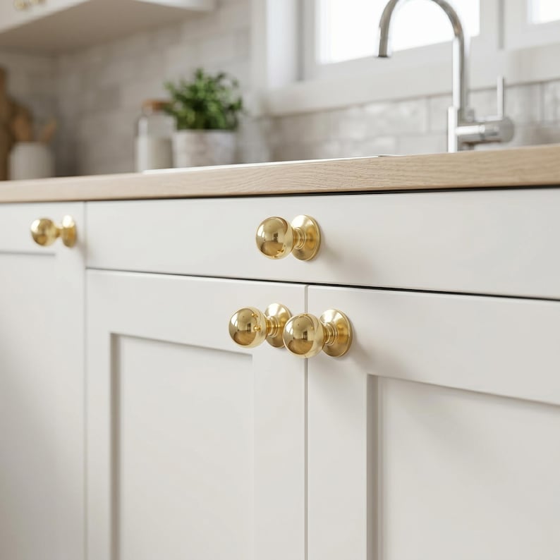 May include: Close-up of white kitchen cabinets with gold-coloured, spherical cabinet knobs. The knobs are mounted on the cabinet doors and drawers. The kitchen has a light wood worktop and a stainless steel tap.