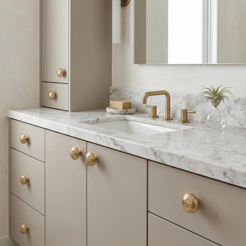 Puede incluir: Un tocador de ba&ntilde;o moderno con encimera de m&aacute;rmol, lavabo blanco y accesorios dorados. El tocador tiene gabinetes de color gris claro con pomos dorados ornamentados. Una peque&ntilde;a planta en un jarr&oacute;n de cristal est&aacute; sobre la encimera.