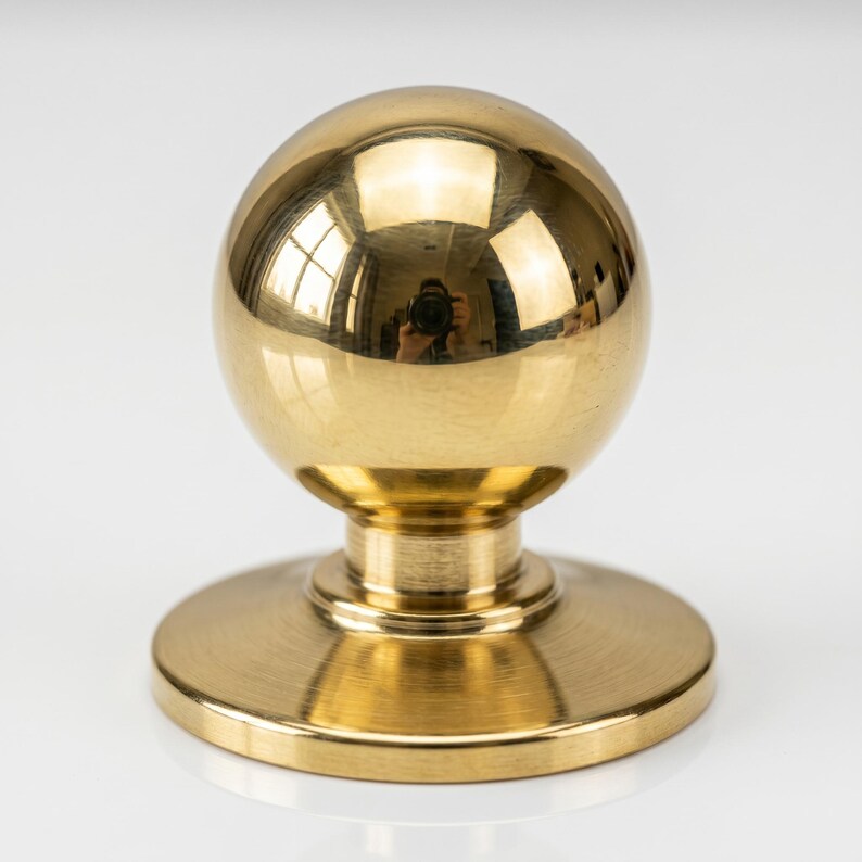 May include: A polished, gold-toned door knob featuring a spherical top and a circular base. The metal surface reflects the surroundings. The knob has a classic design and is made of metal.