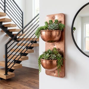 Puede incluir: Una jardinera de pared vertical con dos cuencos de cobre martillado unidos a una placa rectangular de cobre martillado. Los cuencos están llenos de varias plantas suculentas verdes, incluyendo variedades colgantes sobre los bordes.