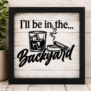 May include: A square framed sign with a white wood-look background and black text that reads I'll be in the... Backyard. The design features a black graphic of a glass of whiskey with ice cubes, a smoking cigar, and an ashtray in the center.