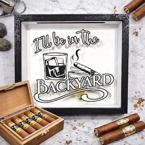 May include: A black framed shadow box with the text "I'll be in the BACKYARD" and a graphic of a whiskey glass and cigar. A wooden cigar box with cigars and loose cigars are also in the image.