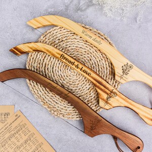 May include: Three wooden bread knives with curved blades and handles. The knives have engraved text that reads "Warm daily", "Bread & Love", and "I am worthy". Each knife features a floral design and a leather loop.