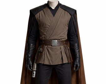 Peut inclure: Un costume marron et noir avec une cape, une ceinture et des bottes. Le costume comprend une tunique marron, un pantalon noir et des gants. Un sabre laser argenté est attaché à la ceinture. Le costume est présenté sur un mannequin.