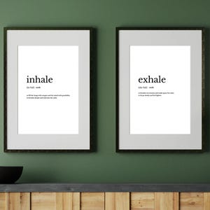 May include: Two framed art prints with black frames hang on a green wall. The left print says "inhale" with a definition. The right print says "exhale" with a definition. Below the prints is a wooden cabinet.