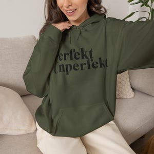 May include: An olive green hooded sweatshirt featuring a front kangaroo pocket, long sleeves, and a drawstring hood. The chest displays the black text "perfekt Unperfekt" in a serif font. The garment is made of a solid-colored fabric.