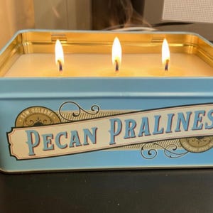 May include: A lit candle in a rectangular, light blue tin with gold trim. The label reads "Pecan Pralines" in a vintage style. Three wicks are lit, and smoke rises from the flames.